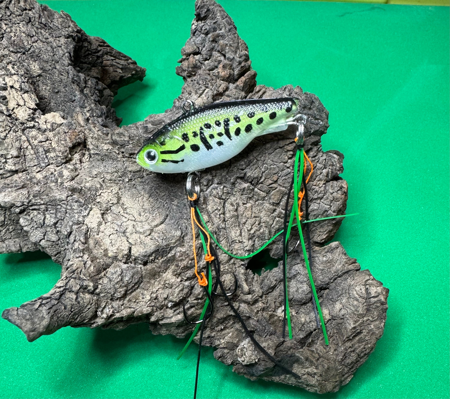 Fishing lure with green, black, and white coloration, positioned on a green surface next to a brown, textured object.