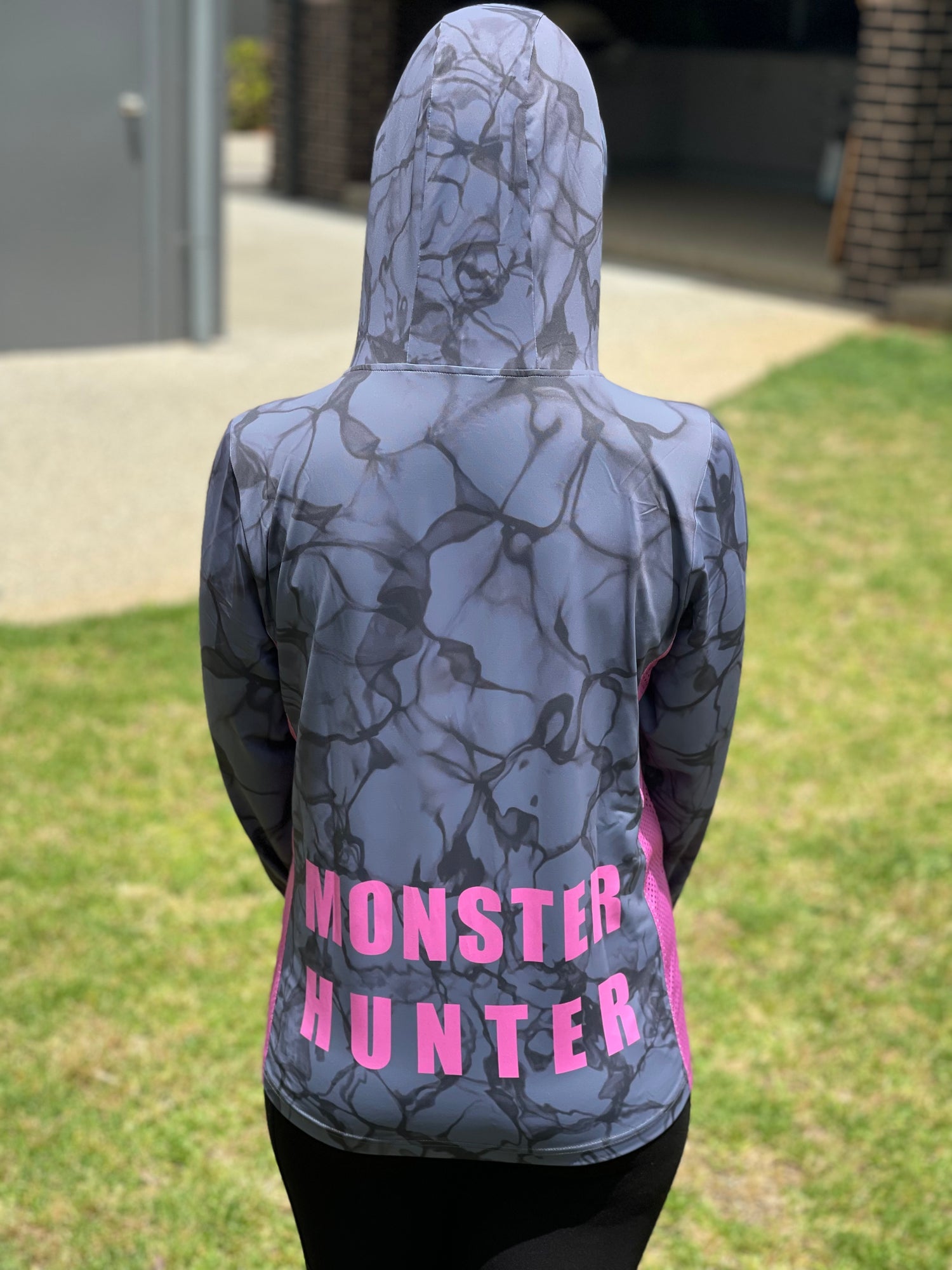 A woman wearing a long-sleeved fishing shirt with a hood, in a camouflage pattern with pink and grey colours, featuring a writing on the back