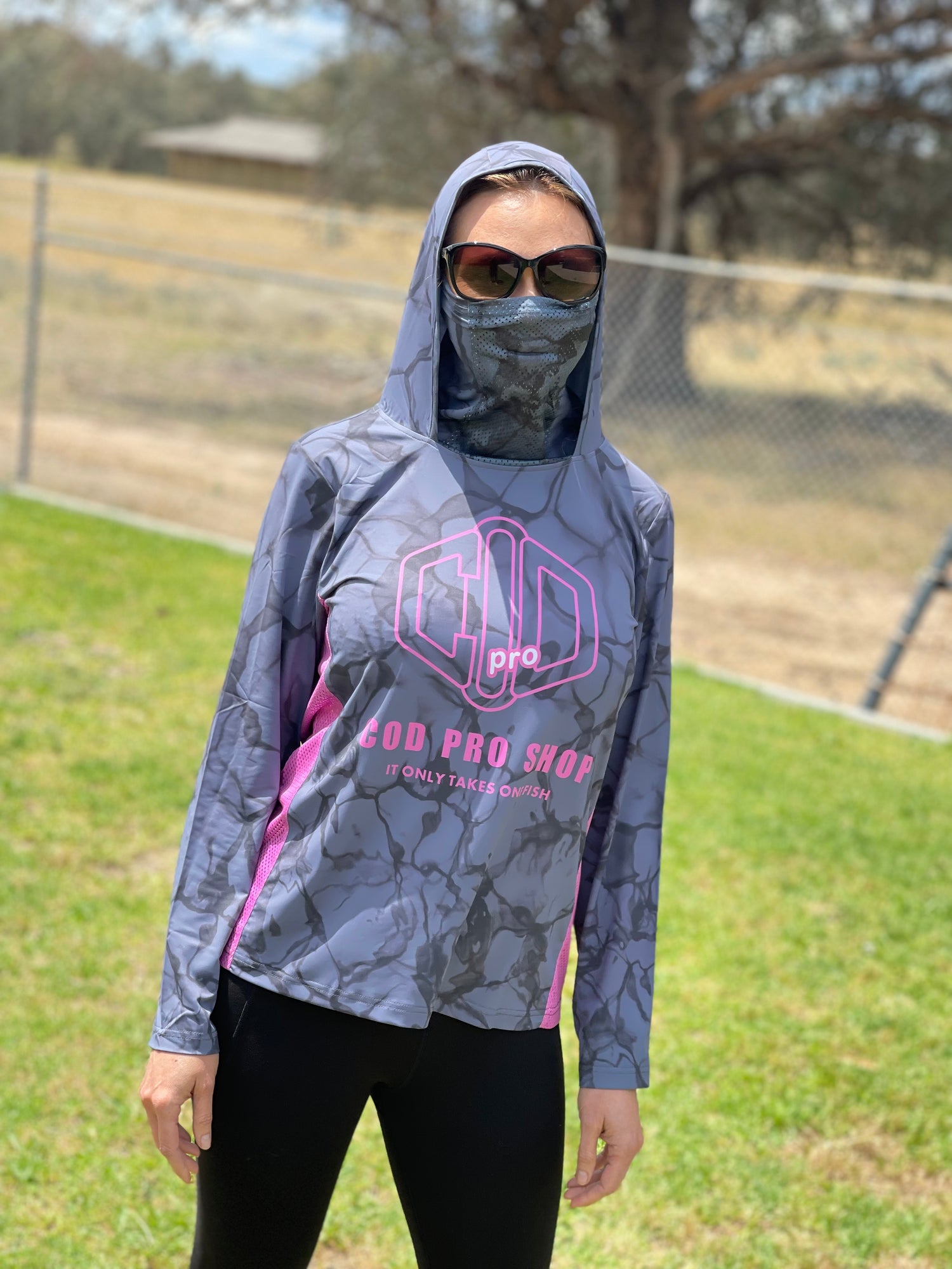 A woman wearing a long-sleeved fishing shirt with a hood, in a camouflage pattern with pink and grey colours, featuring a logo on the chest.