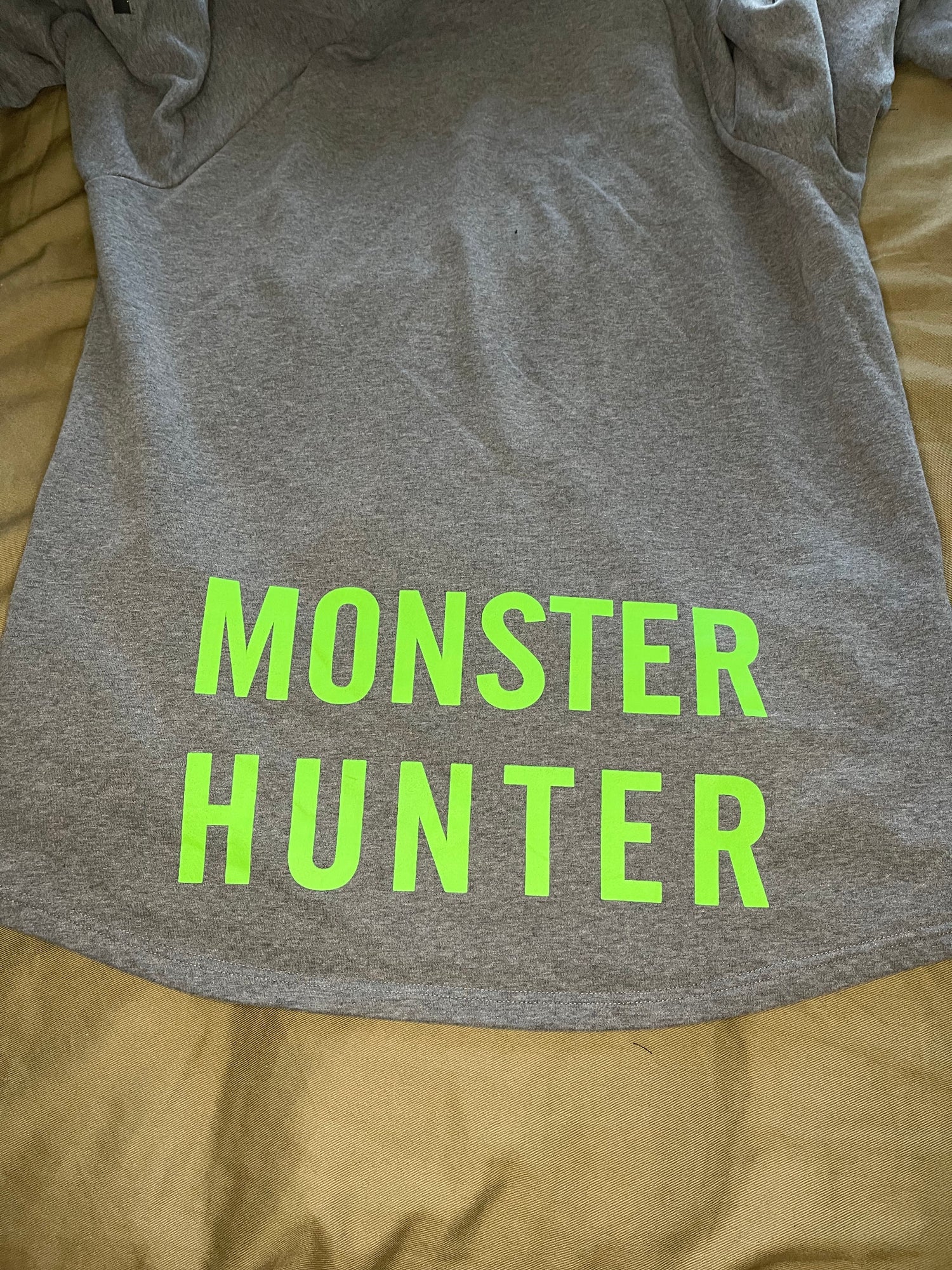 A grey hoodie with the text 'monster hunter' displayed prominently, featuring a hood and a built-in neck scarf. The hoodie is presented on a mannequin against a black background.