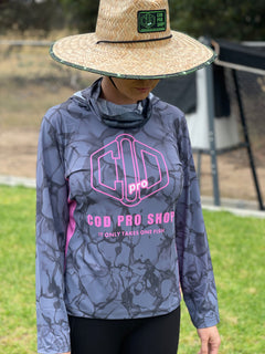 A woman wearing a long-sleeved fishing shirt with a hood, in a camouflage pattern with pink and grey colours, featuring a logo on the chest.
